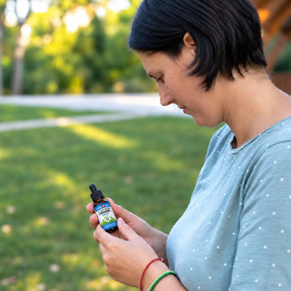 A woman outdoors in a park setting, holding and reading a small bottle of CBD oil. She is wearing a light blue polka-dot t-shirt and red and green bracelets.